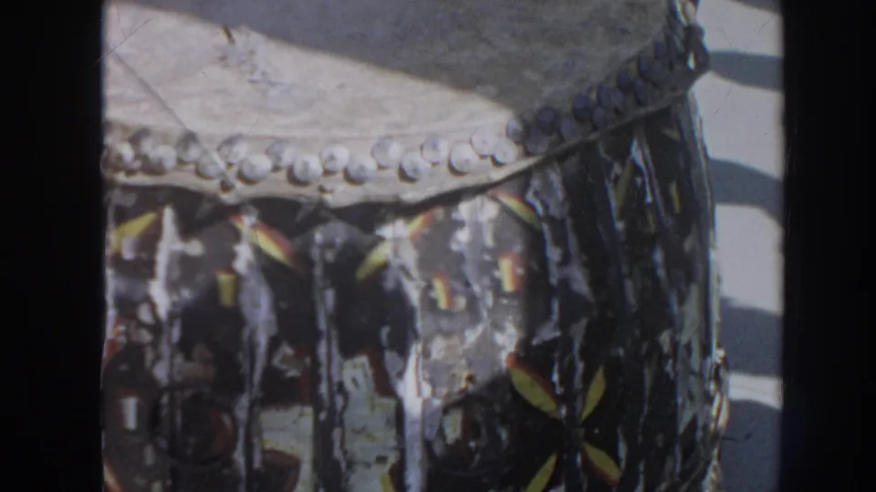Close 1961 Hawaii frame of a decorated drum with textured surface and patterned hanging material in sunlight.