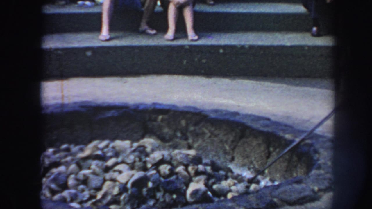 1961 Hawaii 8mm frame showing men at a deep stone-lined fire pit as spectators sit on stepped seating in daylight.