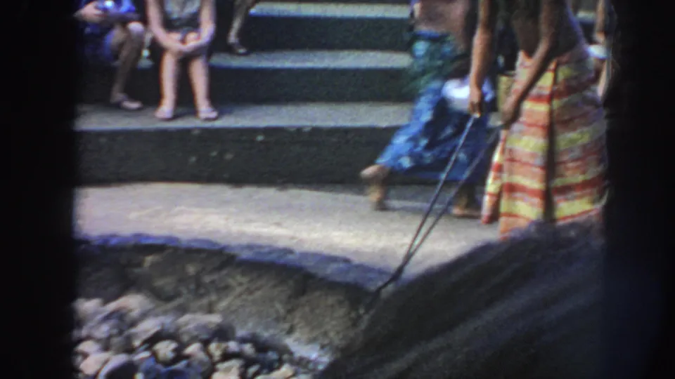 1961 Hawaii frame showing a man in a striped wrap beside the fire pit, with long tools crossing the foreground and spectators on steps behind.