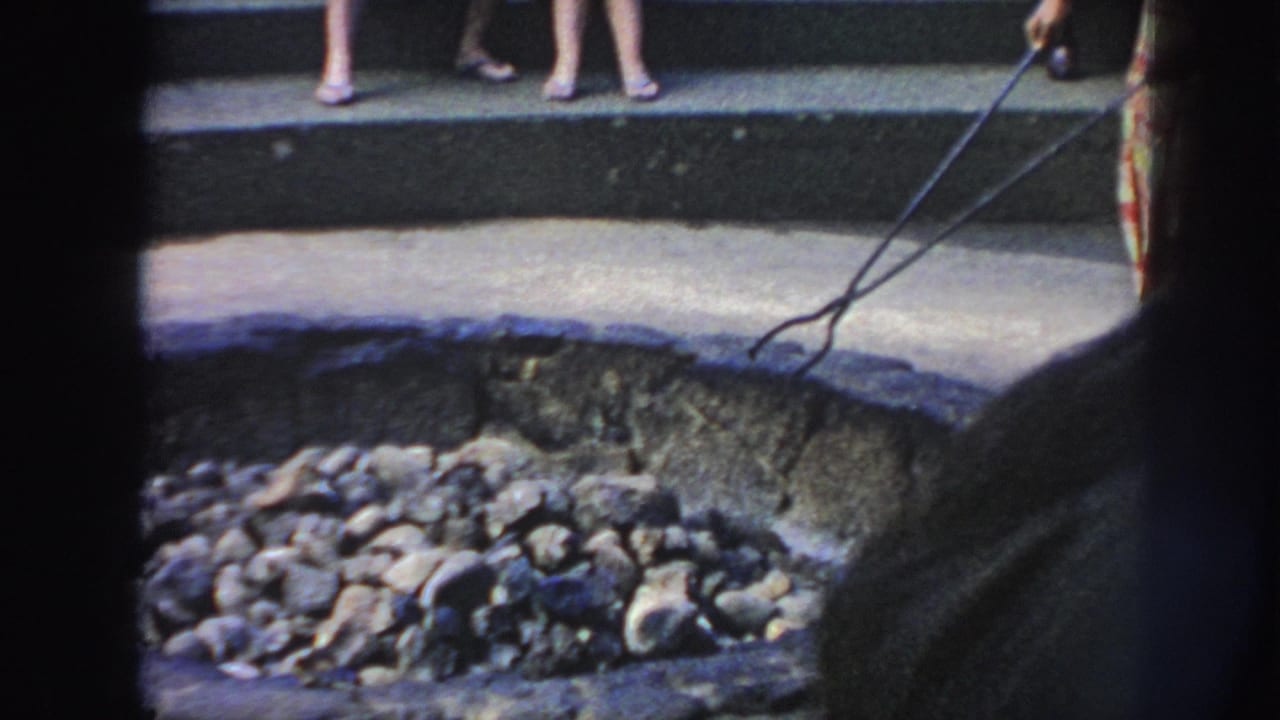 1961 Hawaii 8mm frame showing hot coals in a shallow outdoor pit, with a person holding long tongs and bystanders on steps.