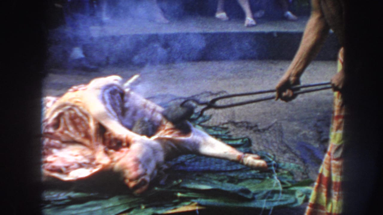 1961 Hawaii frame showing an animal carcass laid on green leaves as smoke rises and long tongs move near the meat.