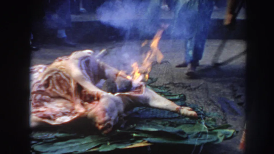 1961 Hawaii 8mm frame showing flames and smoke over an animal carcass on green leaves during outdoor cooking preparation.