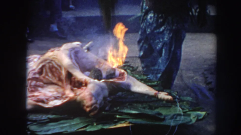 1961 Hawaii frame showing a person beside a smoking, flaming animal carcass on green leaves, with steps and bystanders behind.