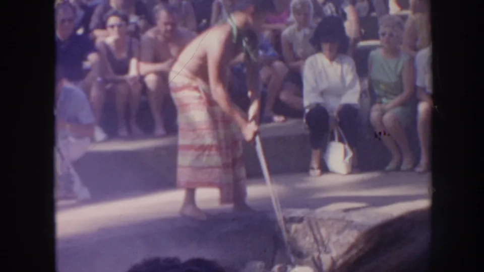 1961 Hawaii frame showing a performer bending with a long-handled tool during a smoke or fire-making demonstration.