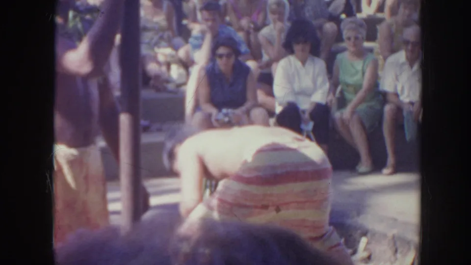 1961 Hawaii 8mm frame showing seated spectators watching a performer or participant in striped clothing at an outdoor gathering.