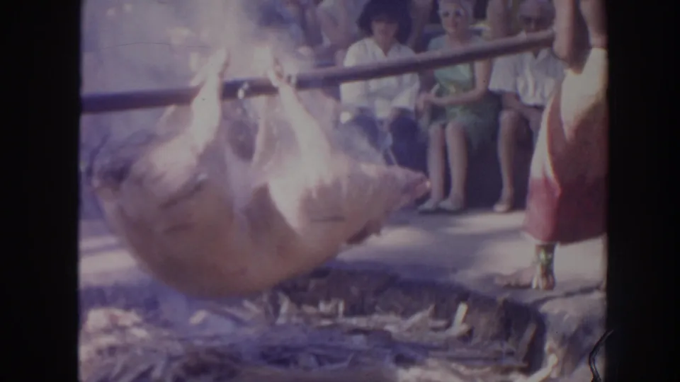 The home-movie frame shows the roasted pig suspended over the pit, with smoke and seated spectators visible behind it.