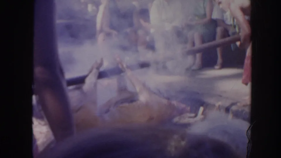 A smoky 1961 Hawaii home-movie frame shows the pig lowered near the pit while spectators remain visible in the background.