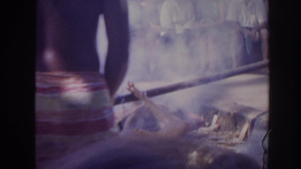 1961 Hawaii 8mm frame showing a whole animal on wood and leaves over smoke, with spectators in the background and a standing figure partly visible.