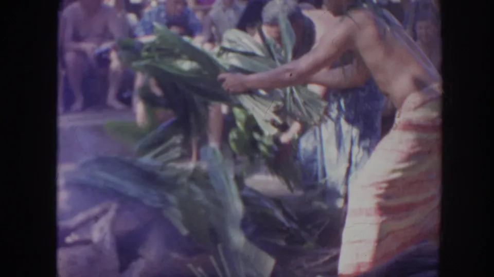1961 Hawaii 8mm frame showing a person in a wraparound skirt tending large green leaves beside the smoky outdoor cooking area.