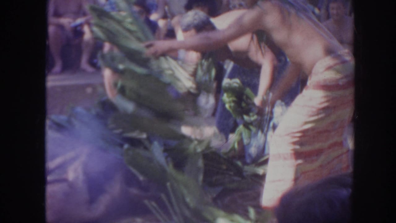 1961 Hawaii 8mm frame of men in wrap skirts and leaf leis arranging banana leaves before a seated audience.