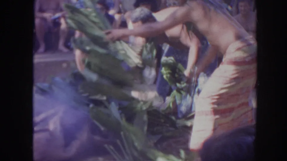 1961 Hawaii 8mm frame of men in wrap skirts and leaf leis arranging banana leaves before a seated audience.