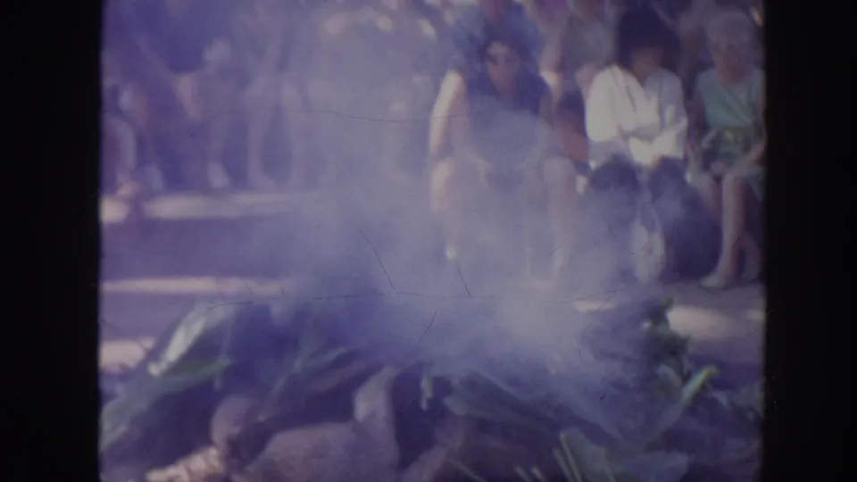 A smoky 1961 Hawaii ceremony frame with banana leaves in the foreground and spectators seated behind.