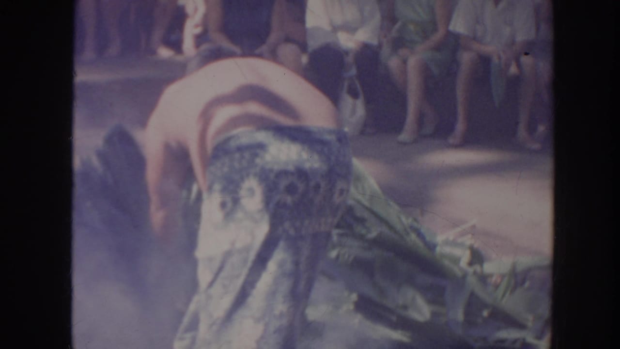 Shirtless performer bends forward near smoke and large leaves with spectators seated behind.