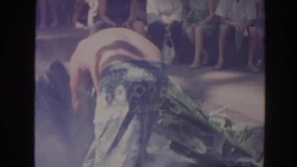 1961 Hawaii 8mm frame of a shirtless performer in a patterned wraparound garment bending near smoke and leaves before an audience.