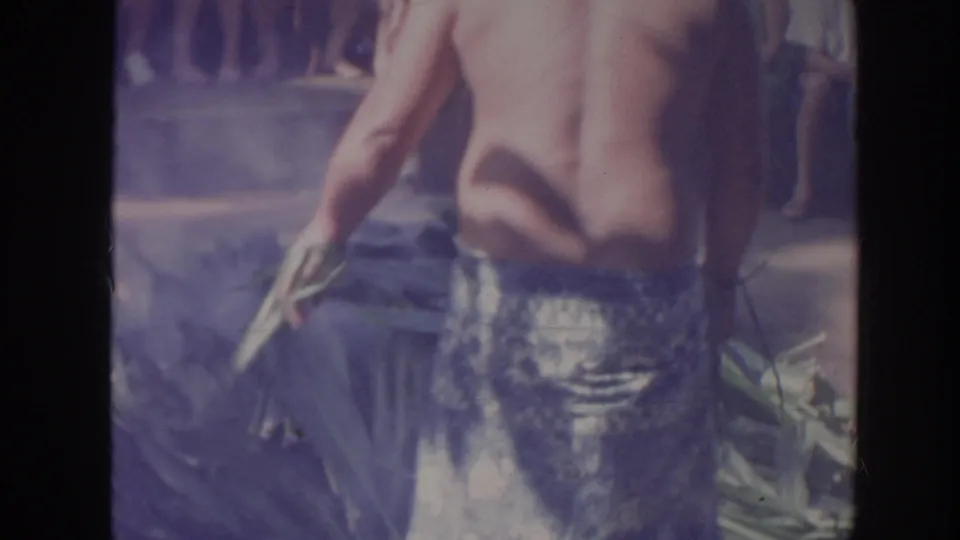 1961 Hawaii 8mm frame showing the back of a shirtless performer in a patterned wraparound skirt beside smoke, leaves, and spectators.