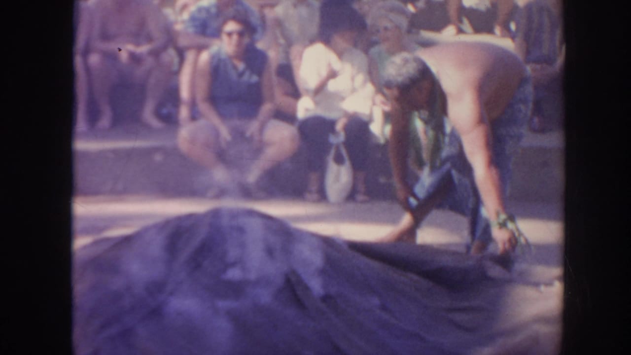 In 1961 Hawaii footage, a shirtless man in a patterned wrap handles a dark pit covering as seated onlookers watch from steps.