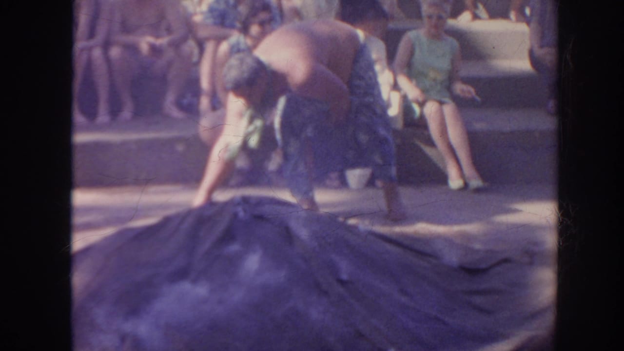 Man reaches across a dark tarp-like covering as seated spectators watch from the background.