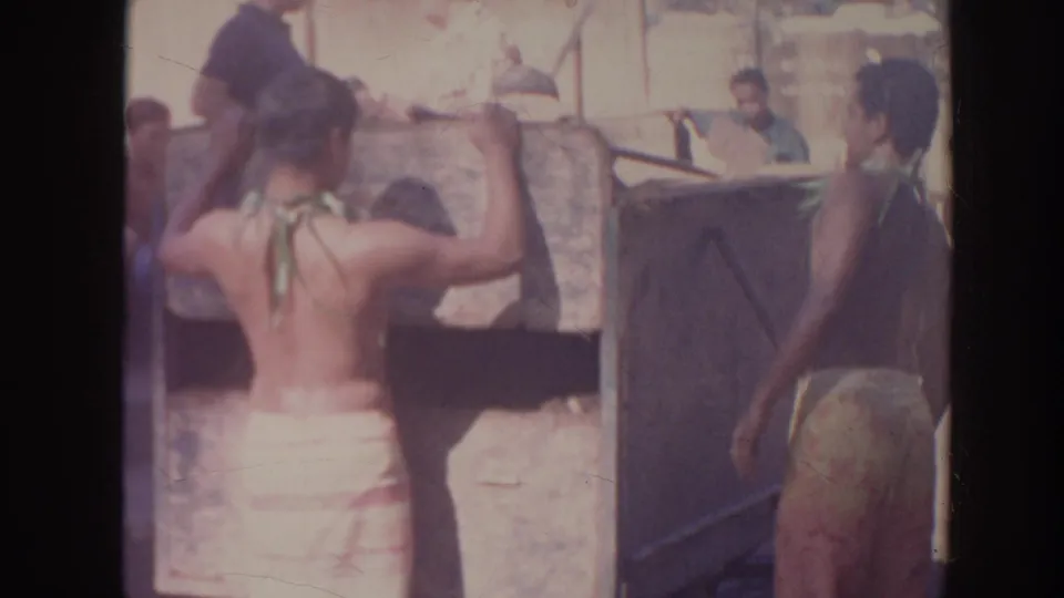Two men in wraps work beside a raised cooking area, with one lifting a dark covering in the 1961 Hawaii home-movie scene.
