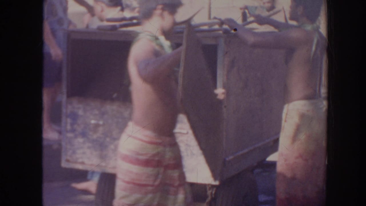In 1961 Hawaii home-movie footage, a shirtless man in a patterned wraparound skirt works with a shovel beside a large wooden box.