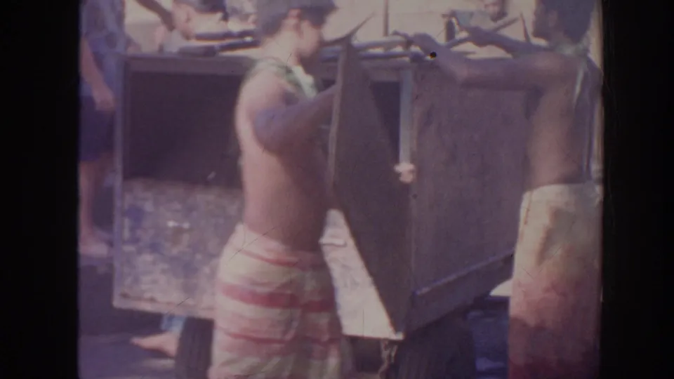 In 1961 Hawaii home-movie footage, a shirtless man in a patterned wraparound skirt works with a shovel beside a large wooden box.