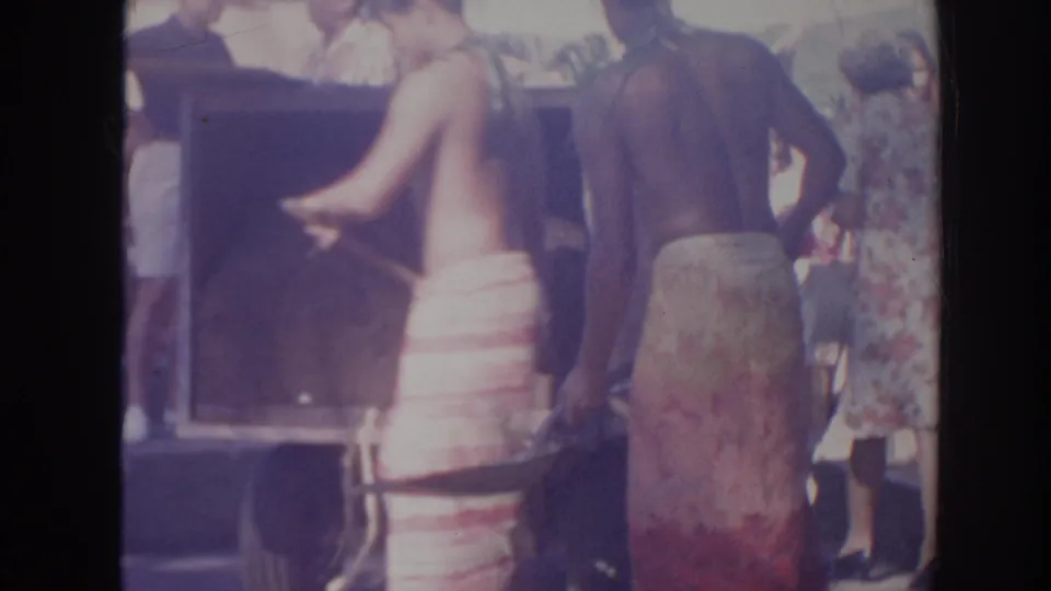 Two shirtless men in wraparound garments stand near a large wooden box in 1961 Hawaii, with shovels and onlookers visible.