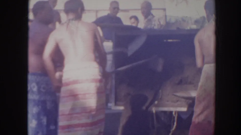 In the 1961 Hawaii home-movie scene, men with shovels work beside a large wooden box as several onlookers stand around them.
