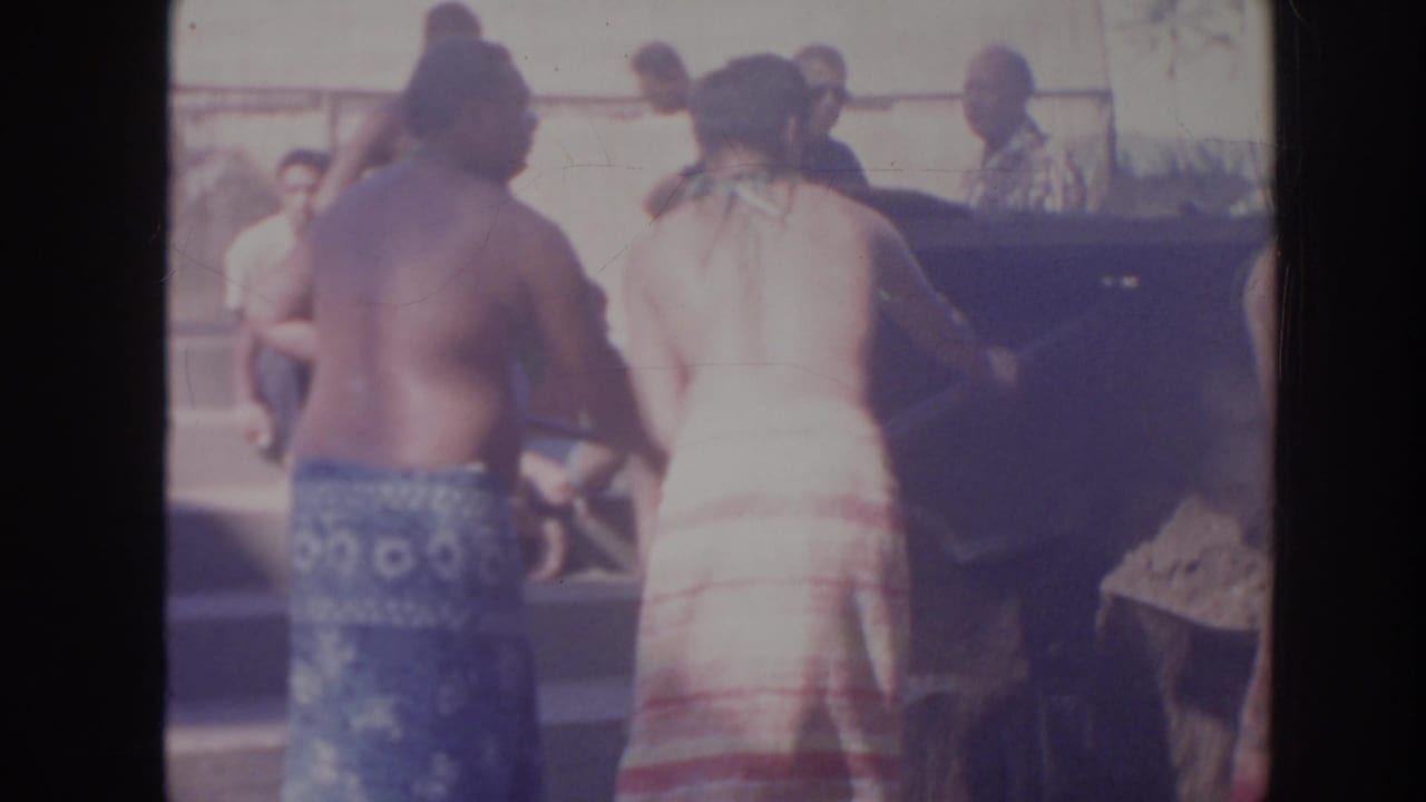1961 Hawaii home-movie frame showing adults, including shirtless men and women in wraparound skirts, gathered near a large object outdoors.