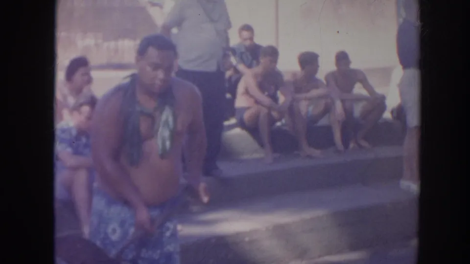 1961 Hawaii frame showing a shirtless man in patterned shorts with seated adults behind him near a large object on concrete.
