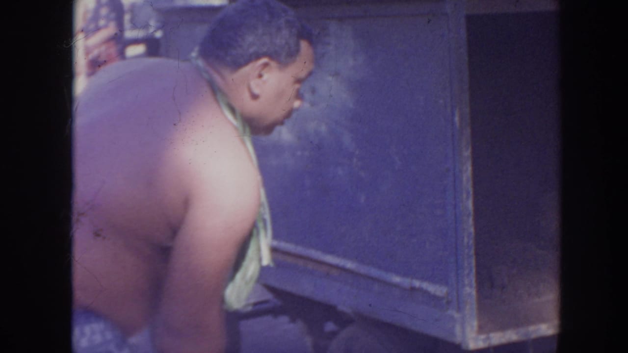 In 1961 Hawaii home-movie footage, a shirtless man wearing a leaf lei leans beside a blue utility box or cooler.