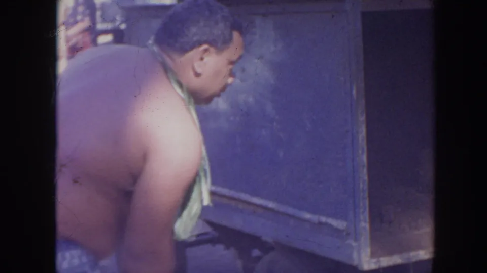 In 1961 Hawaii home-movie footage, a shirtless man wearing a leaf lei leans beside a blue utility box or cooler.