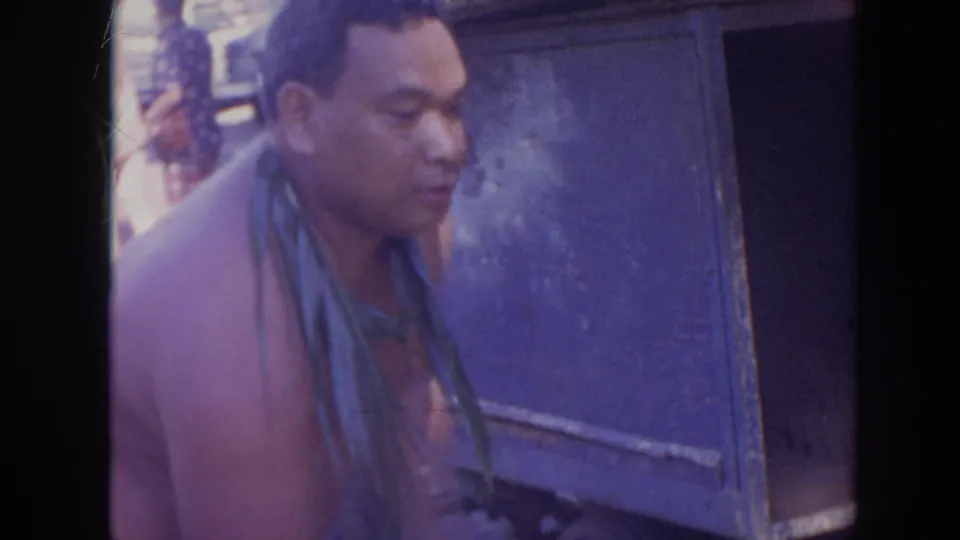 A close 8mm frame shows a man with a leaf lei beside a blue utility box, with faded color and soft focus.