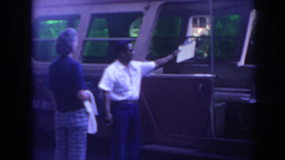 A man in a white shirt holds up a clipboard near a woman and a vintage bus in 1961 Hawaii home-movie footage.