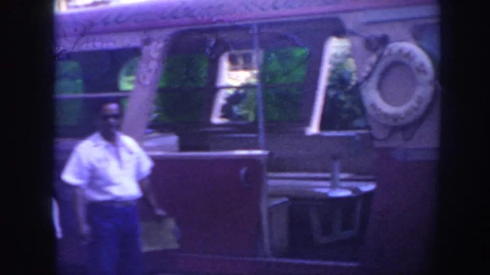 A man stands near the side of a vintage bus with windows and a mounted spare tire, filmed in faded 8mm color.