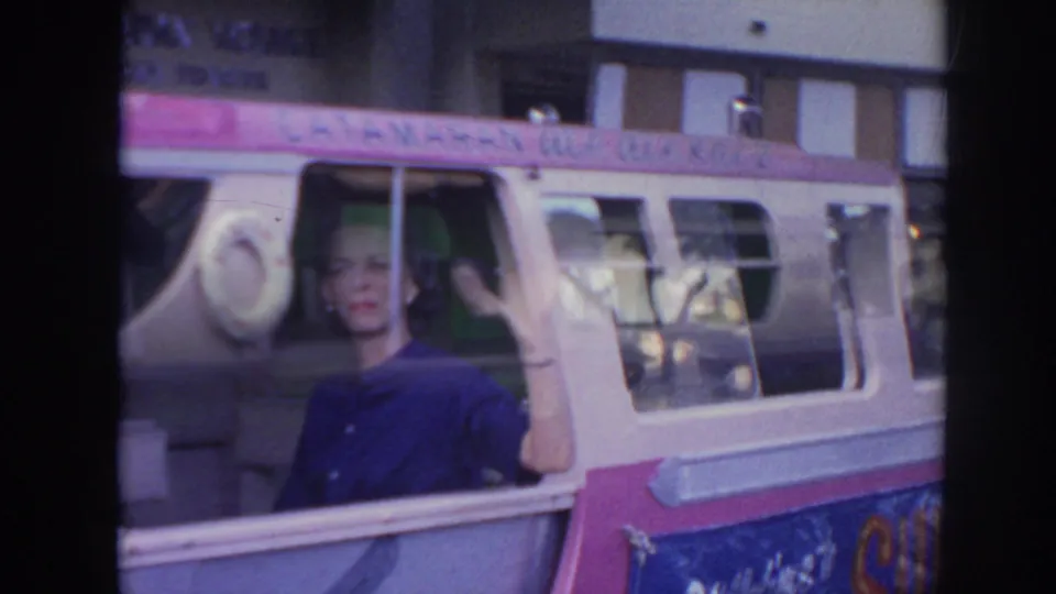 In 1961 Hawaii home-movie footage, a woman smiles and waves from an open window on a pink-and-white tour bus.