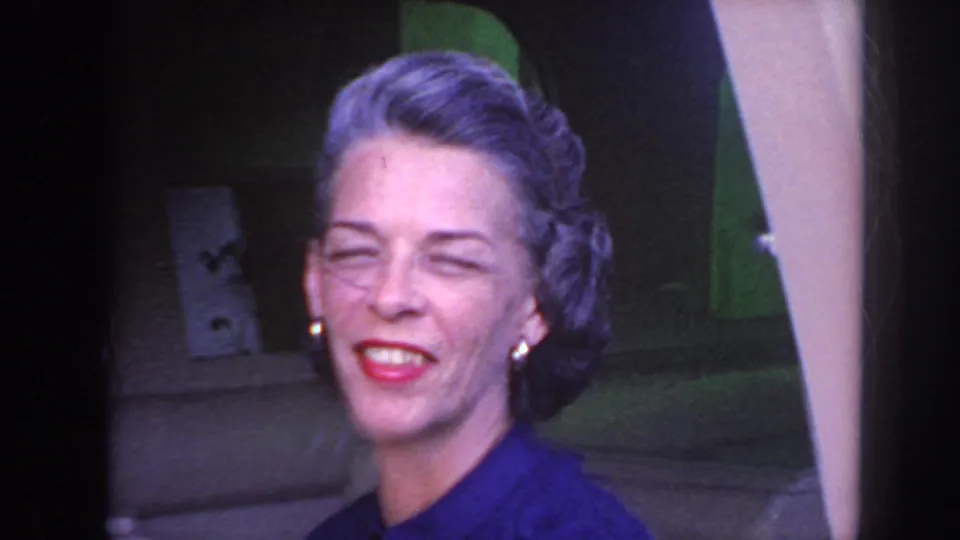 Close 8mm view of the woman smiling near the bus window, with midcentury styling visible in her hair and makeup.