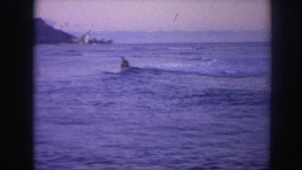 1961 Hawaii home-movie frame showing a distant surfer on calm ocean water beneath a purple-tinged sky and faint shoreline.