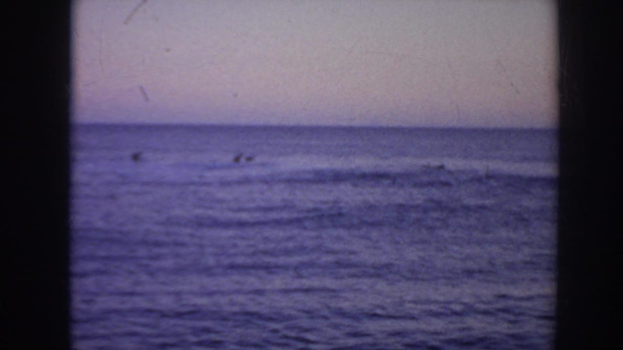 Distant surfers wait beyond small waves under a dim ocean horizon.