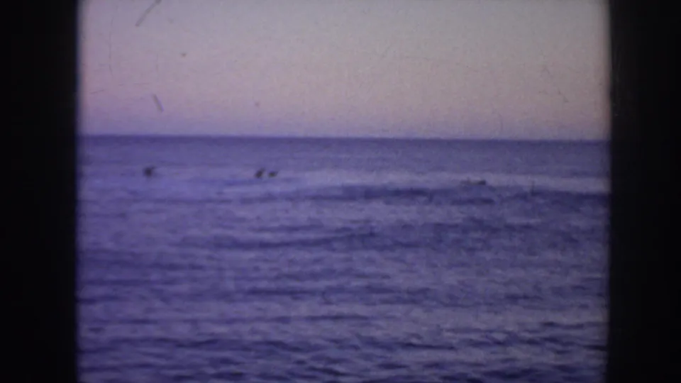 1961 Hawaii 8mm frame showing distant surfers as small silhouettes offshore beneath a muted twilight sky.