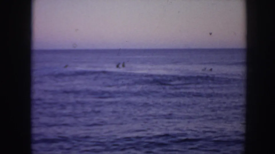 Distant surfers in 1961 Hawaii appear offshore in grainy 8mm footage with a low wave crossing the ocean surface.