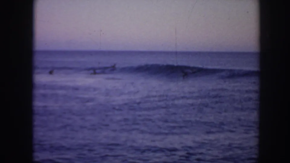 1961 Hawaii home-movie frame with a low breaking wave and distant surfers barely visible against the water.