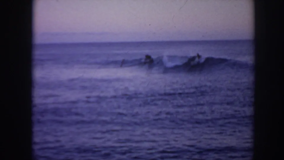 Grainy 1961 Hawaii 8mm frame showing surfers silhouetted near a small breaking wave at dusk.