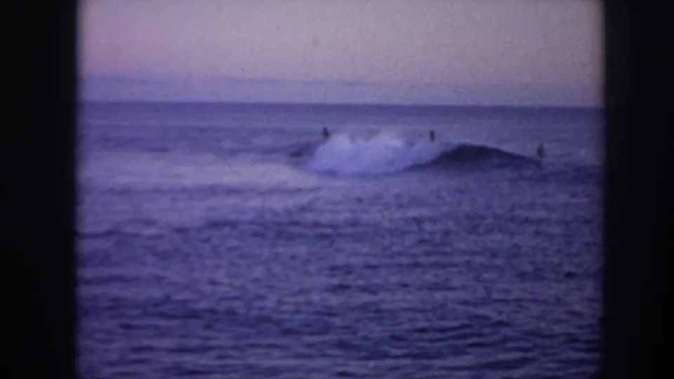 1961 Hawaii 8mm frame showing distant surfers on rolling waves under a hazy blue-purple sky.