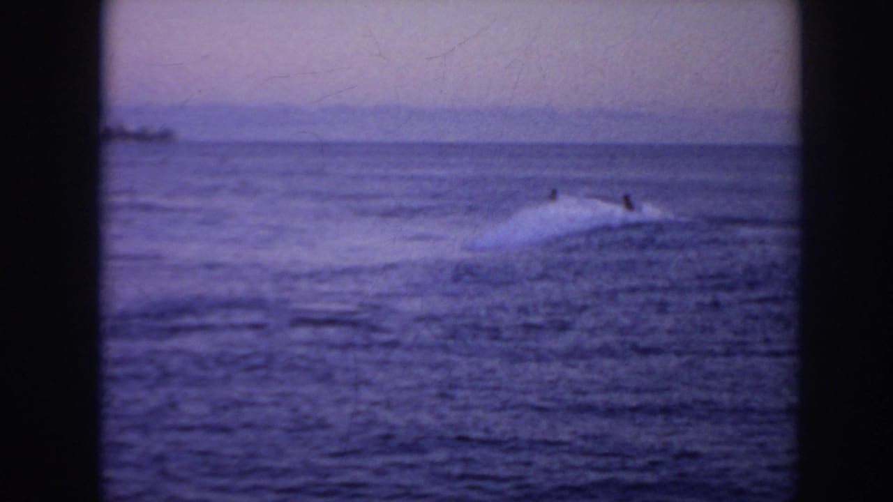 1961 Hawaii home-movie frame with distant surfers beside a low wave on a calm blue-purple sea.