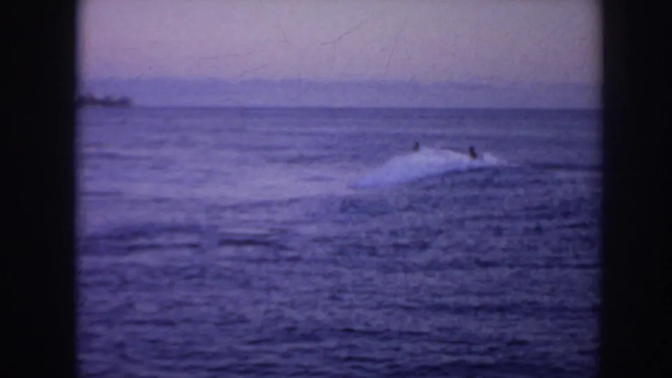 1961 Hawaii home-movie frame with distant surfers beside a low wave on a calm blue-purple sea.