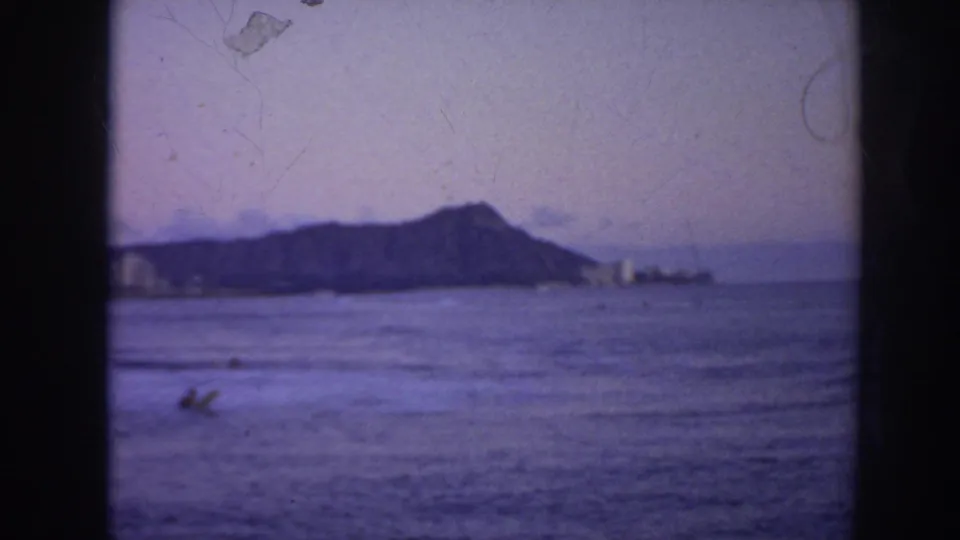 1961 Hawaii home-movie frame showing Diamond Head, distant shoreline buildings, and a surfer low in the foreground water.