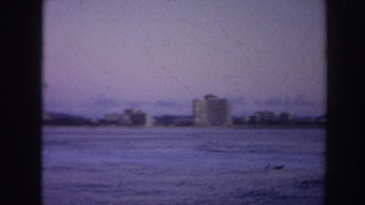 Open water facing a distant shoreline with low buildings and two taller structures.