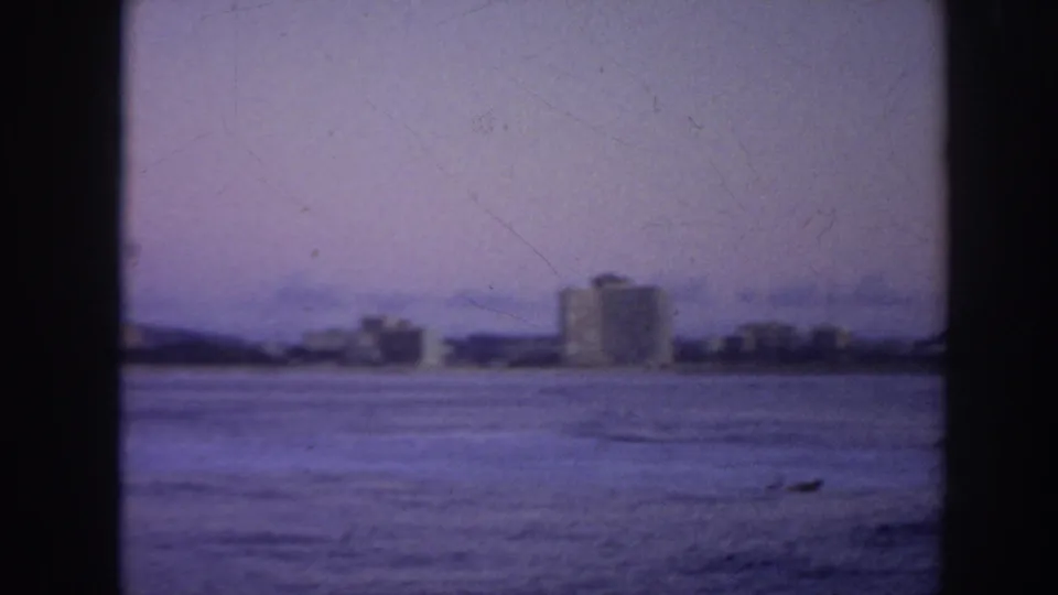 1961 Hawaii coastal view across the ocean toward distant shoreline buildings under a hazy purple sky.