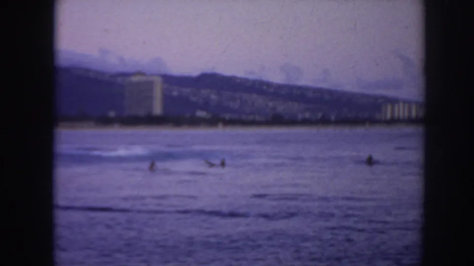 1961 Hawaii coastal frame with small surfers or paddlers in the ocean and hotels along the distant shore.