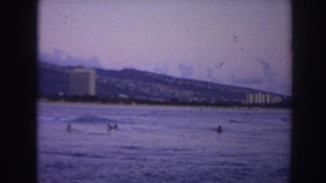 1961 Hawaii home-movie frame showing distant swimmers in the ocean below beachfront buildings and a hazy mountain backdrop.