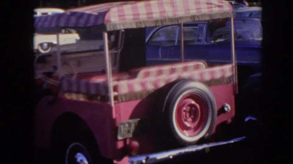 1961 Hawaii frame showing the rear of the pink open vehicle, with red-and-white striped canvas roof, rear bench, and whitewall spare tire.